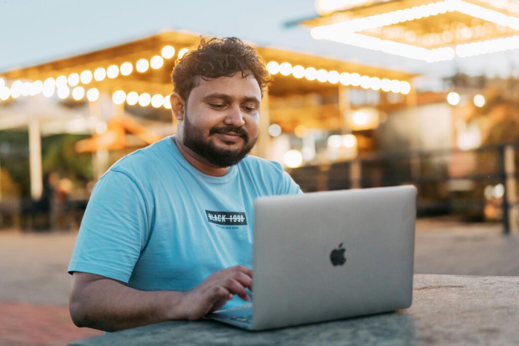 Man smiling and using a laptop while sitting outdoors with lights in the background.